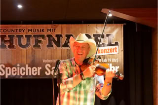 Ein Mann mit Cowboyhut spielt Geige auf einer Bühne, im Hintergrund ein Banner mit der Aufschrift "Country Musik mit Hufnagel".