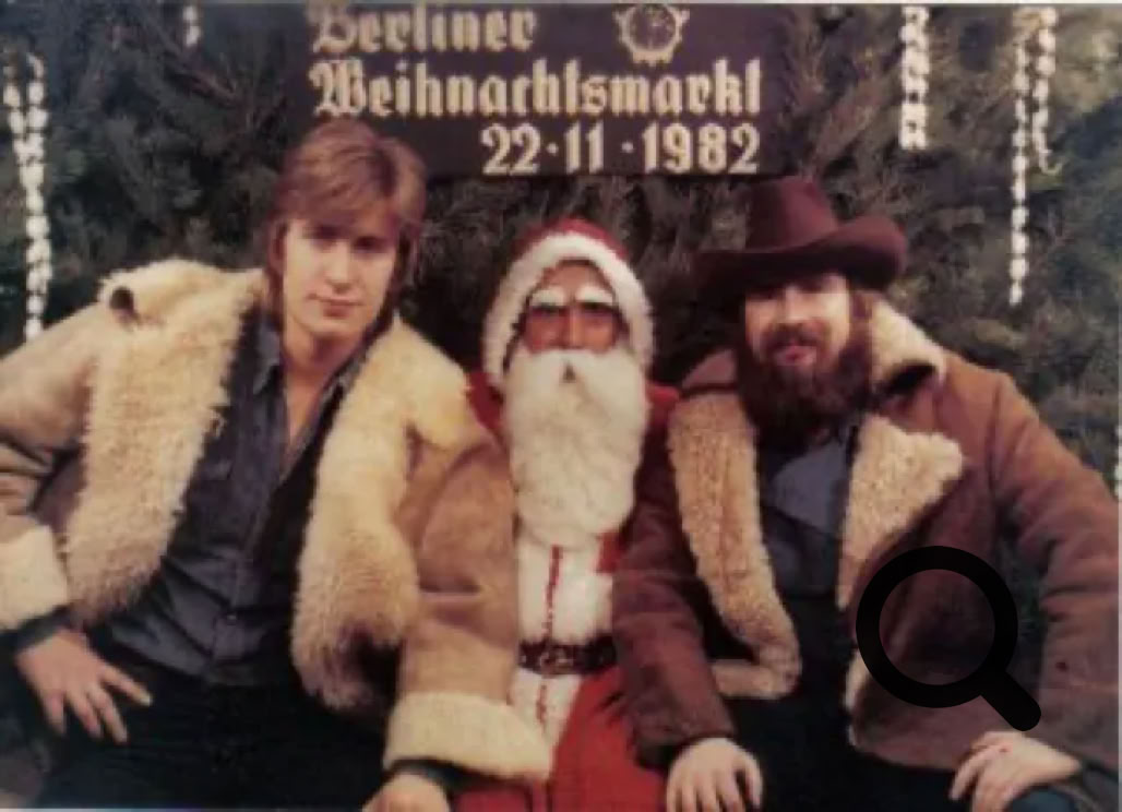 Drei Personen sitzen vor einem Schild mit der Aufschrift "Berliner Weihnachtsmarkt 22-11-1982", in der Mitte eine Person in einem Weihnachtsmannkostüm, die anderen beiden tragen Pelzmäntel.