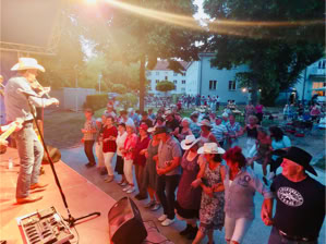 Eine Gruppe von Menschen tanzt vor einer Bühne im Freien, auf der ein Musiker mit Cowboyhut spielt.