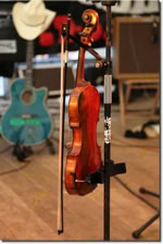 Eine Geige und ein Bogen stehen auf einem Ständer, im Hintergrund sind eine blaue Gitarre und ein weißer Hut zu sehen.