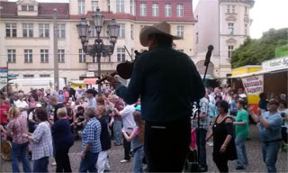 Eine Person mit Hut spielt Geige auf einer Bühne vor einer Menschenmenge auf einem Platz mit historischen Gebäuden im Hintergrund.