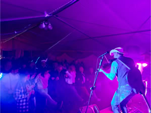 Ein Musiker mit Hut spielt Gitarre auf einer Bühne vor einem Publikum in einem Zelt, beleuchtet von bunten Lichtern.