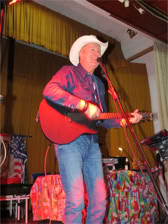 Ein Mann mit weißem Cowboyhut spielt Gitarre und singt auf einer Bühne, umgeben von bunten Vorhängen und Dekorationen.