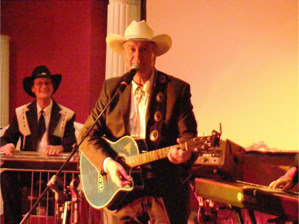 Ein Mann mit Cowboyhut spielt Gitarre und singt, während ein anderer Mann im Hintergrund ein Musikinstrument spielt.