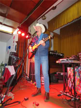 Eine Person mit Cowboyhut spielt Gitarre auf einer Bühne mit rotem Teppich, umgeben von Musikinstrumenten und elektronischen Geräten.