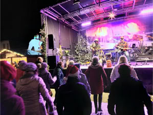 Menschenmenge steht vor einer beleuchteten Bühne mit Musikern, umgeben von Weihnachtsdekorationen und Bäumen.