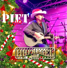 Eine Person mit Cowboyhut spielt Gitarre auf einer Bühne, umgeben von Weihnachtsdekorationen und einem Schild mit der Aufschrift "Hüttengaudi".