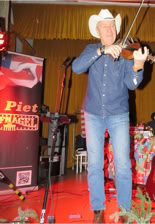 Ein Mann in Jeans und Cowboyhut spielt Geige auf einer Bühne, im Hintergrund ein Banner und ein gelber Vorhang.